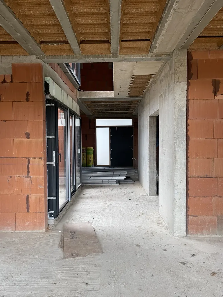 Passage intérieur en chantier avec murs en briques, poutres en béton et menuiseries extérieures