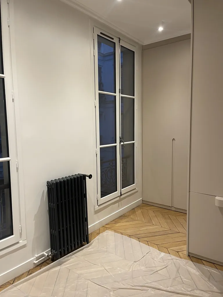 Pièce avec fenêtre blanche, radiateur noir, placard et parquet en chevrons partiellement protégé